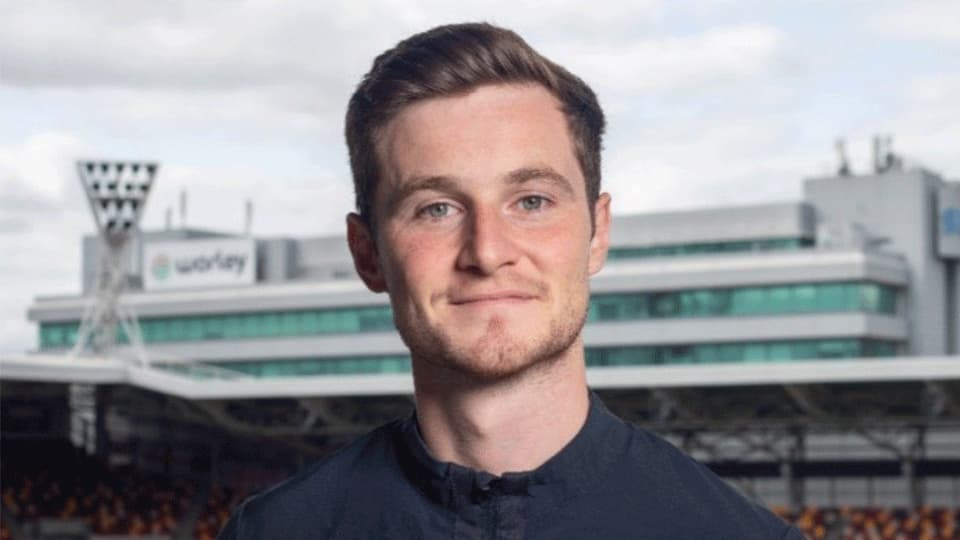A headshot of Joe Jackson wearing a navy top with the Brentford Football Club logo patch on the the Umbro logo in white. He is in focus standing in front of a football pitch and stadium which is blurred in the background.